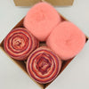 Three balls of yarn in a brown gift box: two variegated yarns in red, orange, and pink tones, and two fluffy solid pink yarns. A partial brown box lid is visible at the top against a white background.