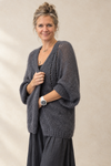 A woman wearing a chunky, open-knit charcoal gray cardigan over a black top and dark skirt, standing against a neutral beige textured wall.